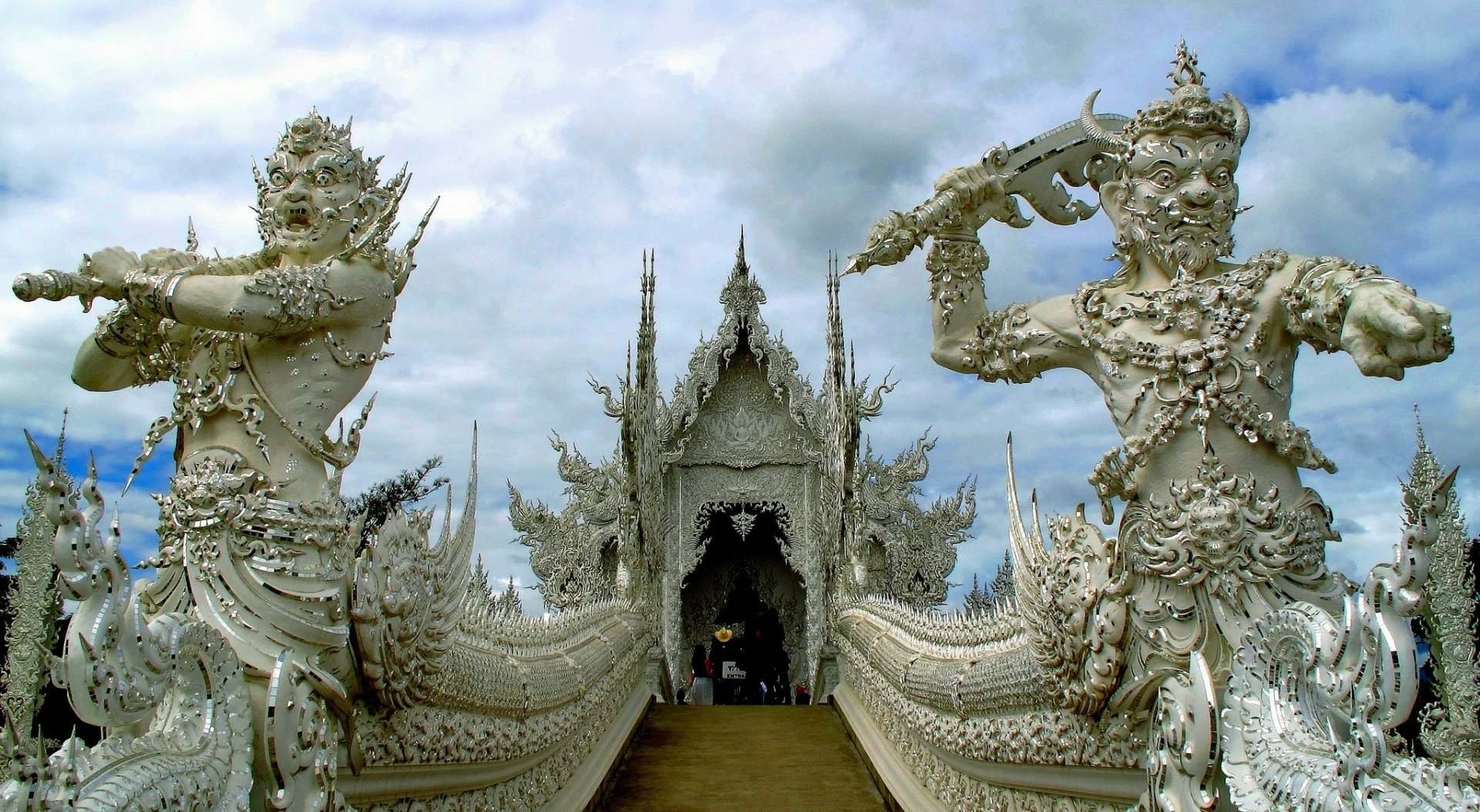 A stunning view of Wat Rong Khun, a remarkable white temple in Chiang Rai Province, Thailand. Intricate sculptures guard the entrance, set against a dramatic sky.