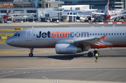 An Airbus A320 Jetstar aircraft is shown taxiing at Sydney Airport, with a ground crew member in view, capturing the bustling atmosphere of aviation.