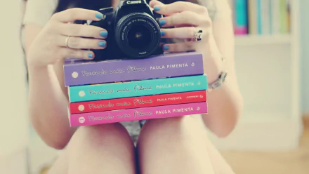 A person holds a Canon camera while resting colorful books on their legs, set against a softly blurred backdrop of bookshelves. This HD image creates a cozy, intellectual atmosphere.