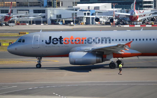 An Airbus A320 Jetstar aircraft is shown taxiing at Sydney Airport, with a ground crew member in view, capturing the bustling atmosphere of aviation.