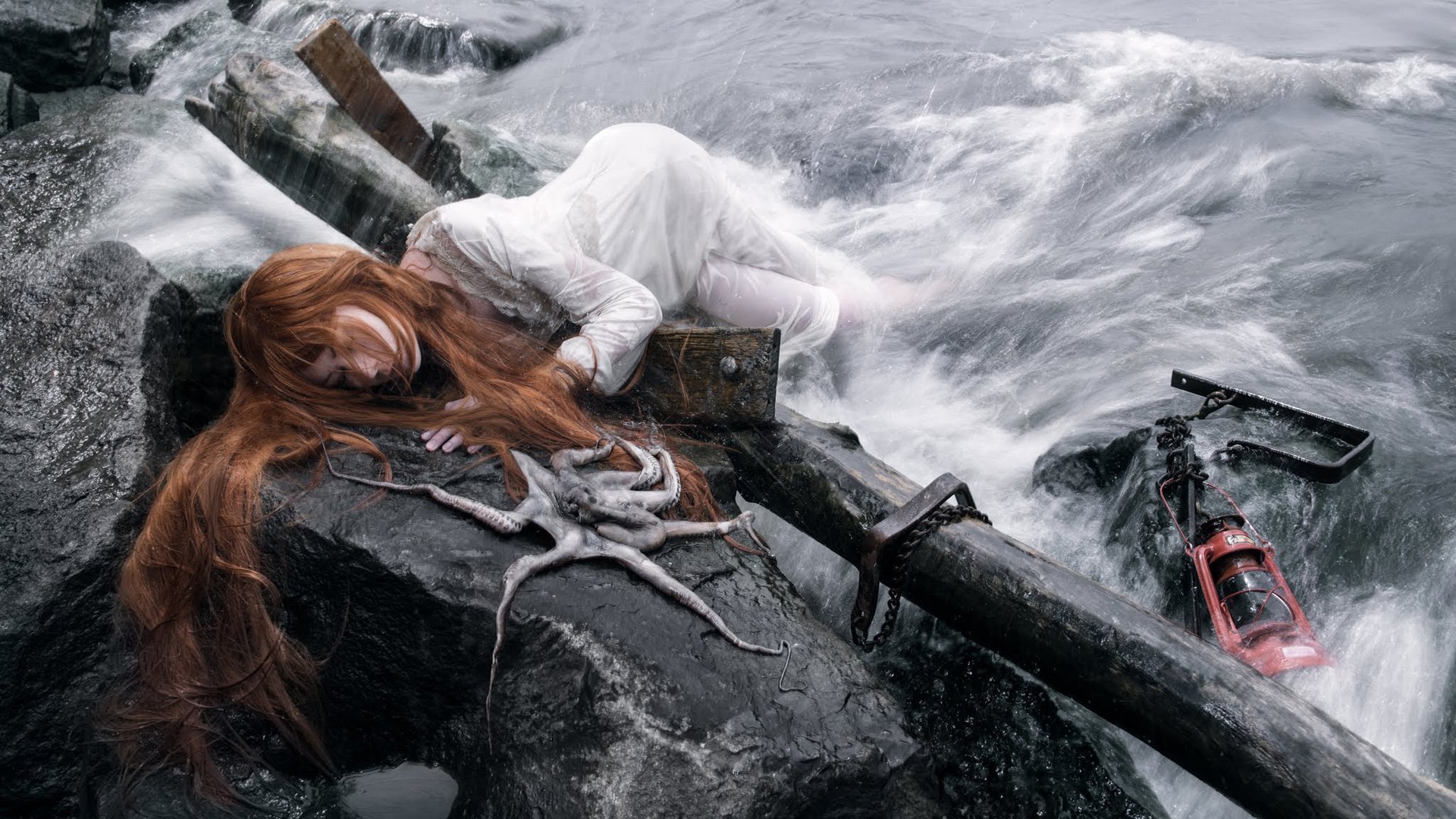 Artistic HD wallpaper featuring a woman in a white dress lying on rocky shores with flowing red hair, surrounded by water. An octopus and various objects are scattered around her.