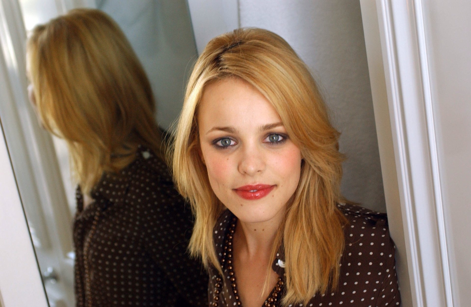 HD PC desktop wallpaper featuring celebrity Rachel McAdams with blonde hair, wearing a dark patterned blouse, posing beside a mirror in a softly lit setting.