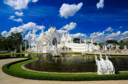 Stunning HD desktop wallpaper of Wat Rong Khun, a white religious temple in Thailand, set against a vibrant blue sky with lush greenery and reflective water.