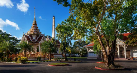 HD PC desktop wallpaper: sunlit Thai temple complex with ornate spired pavilion among trees and a vivid blue sky, showcasing Thailand's religious architecture and tranquil grounds.