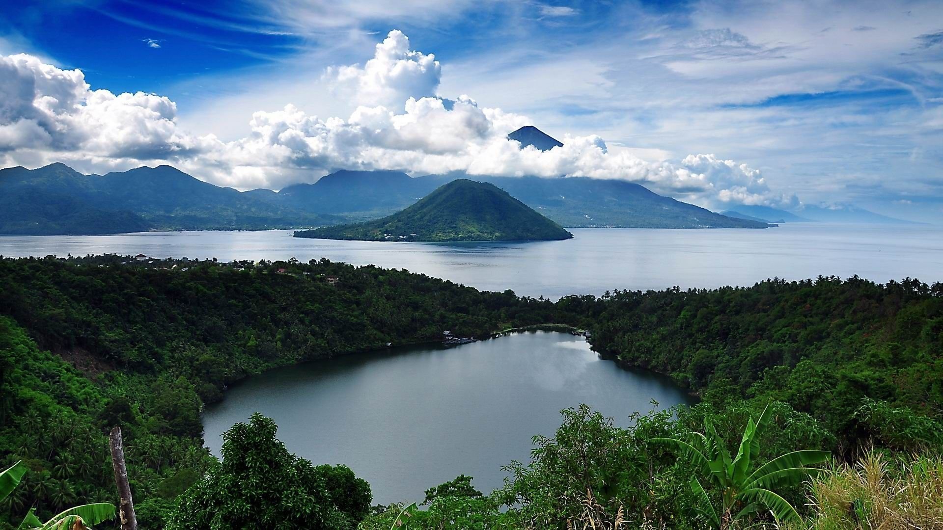 HD desktop wallpaper of a lush green island surrounded by calm water, with mountains and a partly cloudy sky in the background, showcasing vibrant nature scenery.