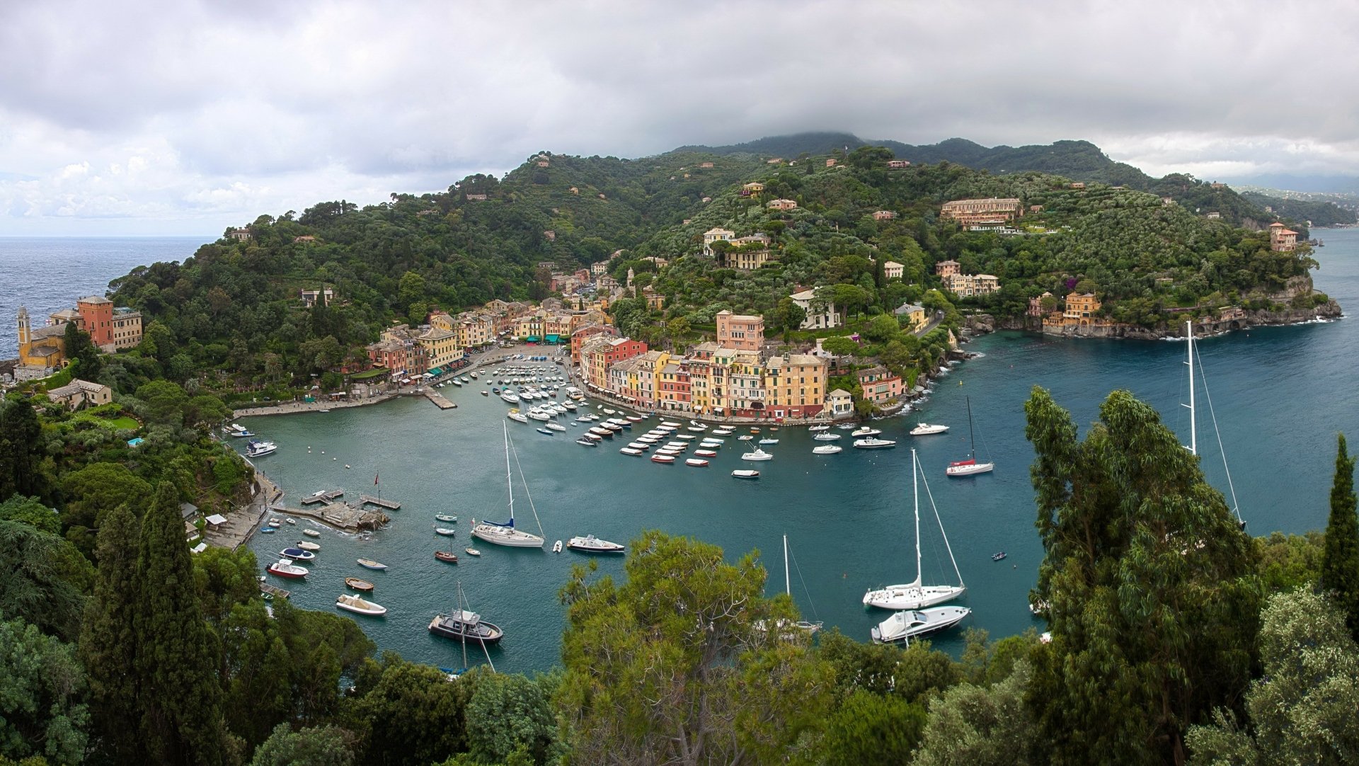 A vibrant coastal village in Portofino, Liguria, Italy, featuring colorful houses, boats docked in the water, lush forested hills, and a scenic island backdrop.