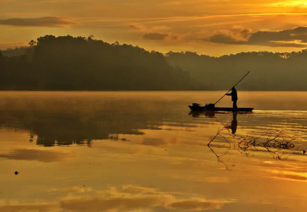 HD photography desktop wallpaper showing a person on a small boat fishing on a calm lake at sunset with silhouetted hills and warm golden reflections on the water.