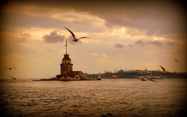 A serene HD wallpaper of Istanbul showcasing the Maiden's Tower amidst a calm sea, with birds soaring above and a dramatic sunset illuminating the skyline.
