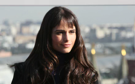 2K Quad HD PC wallpaper: woman with long dark hair and bangs, wearing a dark coat and faint smile, posed against a blurred city skyline background.