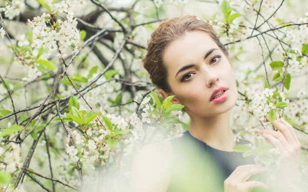 HD PC desktop wallpaper: close-up of a woman's face framed by white blossoms and green leaves, soft-focus background evoking a dreamy spring mood.