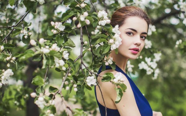 HD desktop wallpaper of a woman in a blue dress among blooming spring branches, highlighting her serene face and natural beauty, captured by Lidia Sergeeva.
