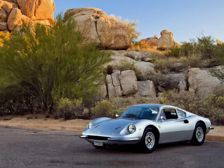 Silver Ferrari Dino 246 GT vehicle parked on a desert road against rocky boulders and shrubs — HD PC desktop wallpaper background.