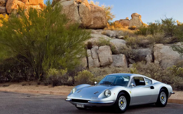 Silver Ferrari Dino 246 GT vehicle parked on a desert road against rocky boulders and shrubs — HD PC desktop wallpaper background.