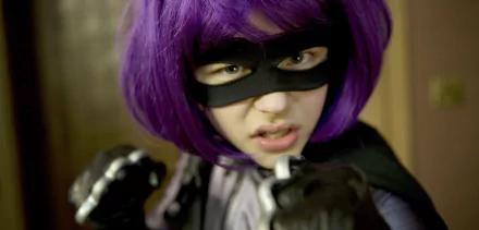 Close-up of masked Hit-Girl in a purple wig and black costume from the Kick-Ass movie, pointing a gloved fist toward the camera — 2K Quad HD PC desktop wallpaper.