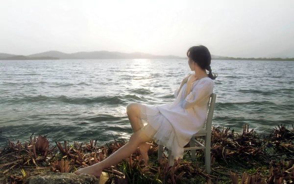 A barefoot woman in a white dress sits by the sea, her legs stretched toward the water, gazing at the serene landscape. The image exudes tranquility and beauty, making it an ideal HD desktop wallpaper.
