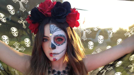 Artistic sugar skull face paint on a person wearing a red-and-black floral crown against a skull-patterned backdrop — HD PC desktop wallpaper and background.