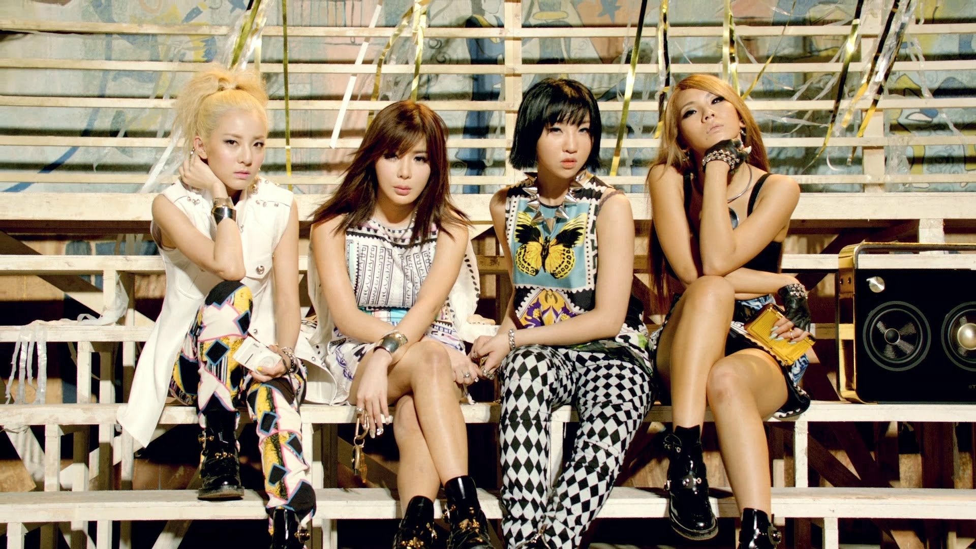 HD PC desktop wallpaper featuring the music group 2NE1 seated on bleachers, showcasing their distinctive fashion and strong stage presence.
