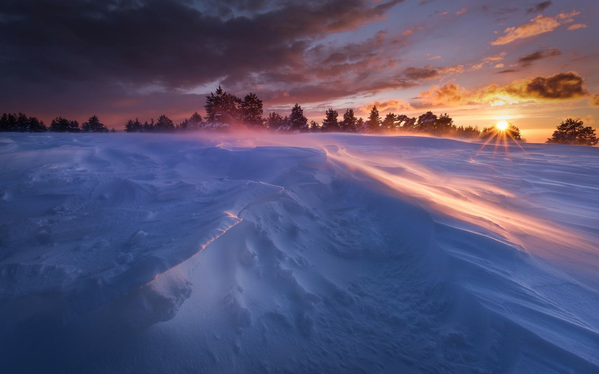 A stunning HD wallpaper showcasing a vibrant sunset over a snowy landscape, with gentle light illuminating the snowdrifts and silhouetted trees against the colorful sky.