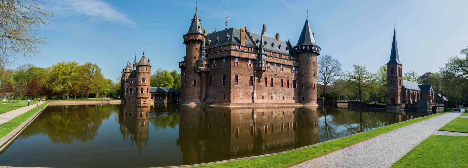 Panoramic view of Castle De Haar in Utrecht, Netherlands, showcasing its reflective moat and historic architecture under a clear blue sky. HD wallpaper and background.