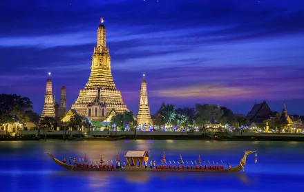 Night view of Wat Arun Temple in Bangkok, Thailand, illuminated along the river with a traditional boat in the foreground against a deep blue sky.