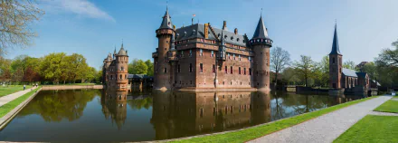 Panoramic view of Castle De Haar in Utrecht, Netherlands, showcasing its reflective moat and historic architecture under a clear blue sky. HD wallpaper and background.