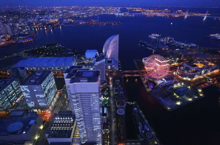Nighttime aerial view of Yokohama, Japan, showcasing illuminated man-made structures and waterfront with vibrant city lights against the dark sky.