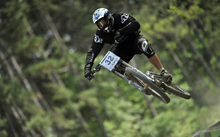 A high-definition desktop wallpaper featuring a BMX rider in mid-air, performing a jump against a blurred forest background. The rider wears a helmet and sports a numbered bib, emphasizing a sporting event.