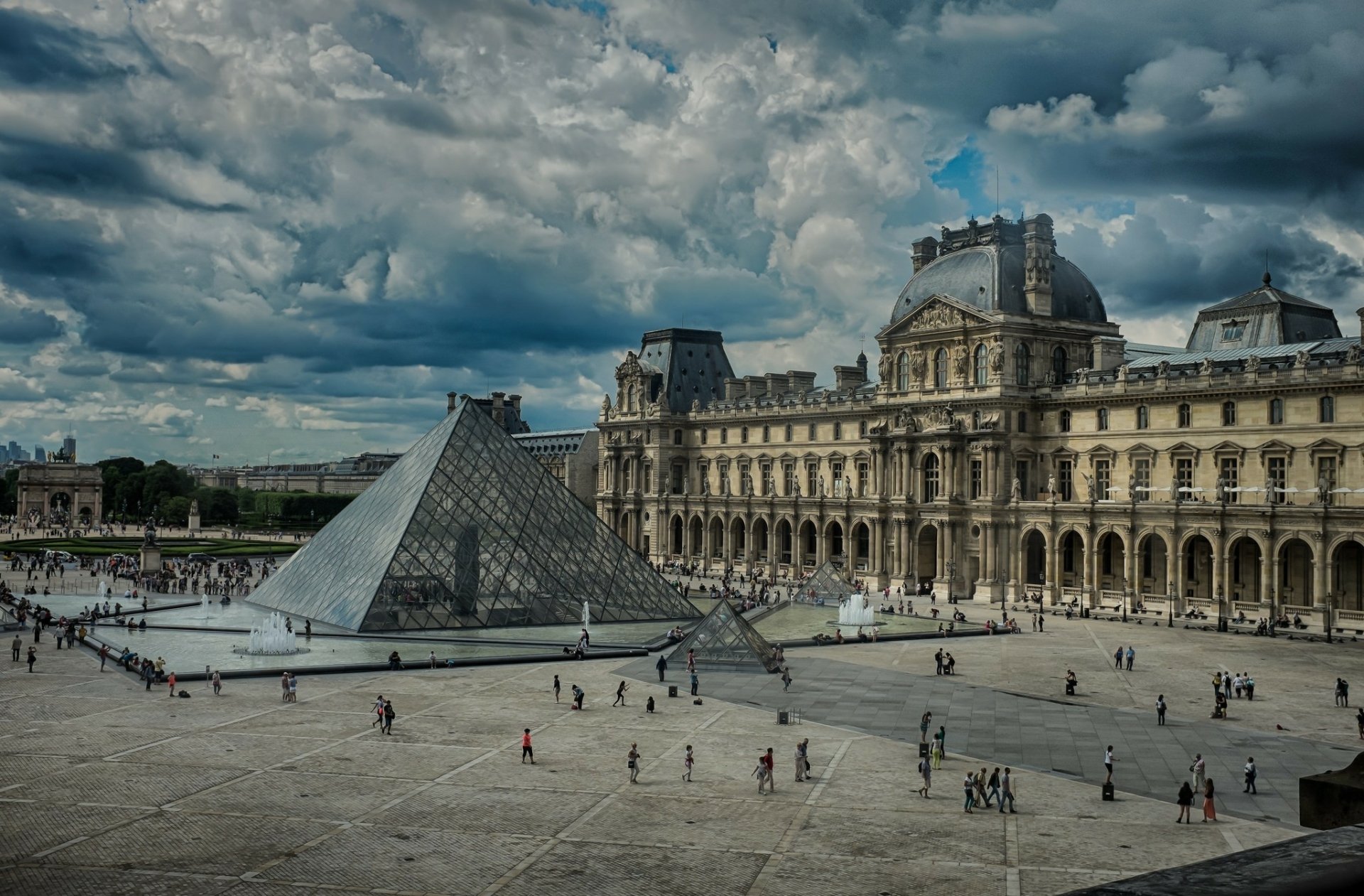 A stunning view of the Louvre in Paris, featuring the iconic glass pyramid against a dramatic cloud-filled sky, showcasing the beauty of man-made architecture in a historic setting.