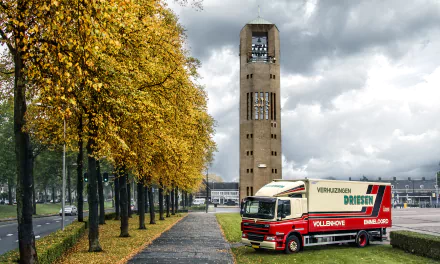 4K Ultra HD image of a tree-lined path with autumn foliage, a truck parked on grass, and a tall bell tower under a cloudy sky in the Netherlands.