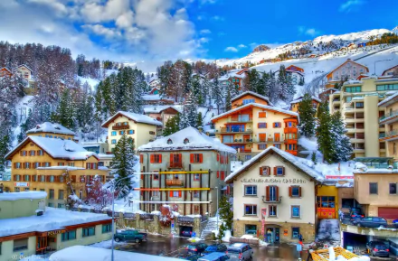 Snow-covered winter village in St. Moritz, Switzerland, showcasing charming man-made buildings against a mountainous, blue-sky backdrop in HD quality.