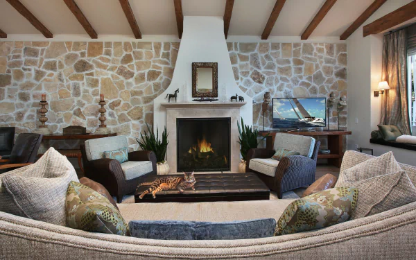 Cozy living room with stone fireplace, TV set, cushioned sofa and chairs, a cat lounging on a rug, soft lighting, and wooden beam ceiling in HD desktop wallpaper style.