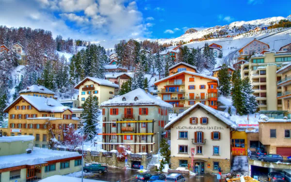 Snow-covered winter village in St. Moritz, Switzerland, showcasing charming man-made buildings against a mountainous, blue-sky backdrop in HD quality.