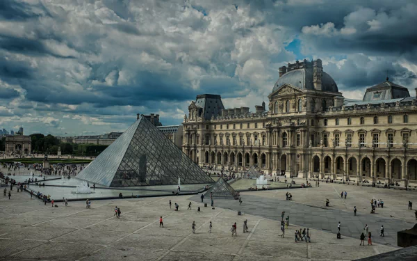 A stunning view of the Louvre in Paris, featuring the iconic glass pyramid against a dramatic cloud-filled sky, showcasing the beauty of man-made architecture in a historic setting.