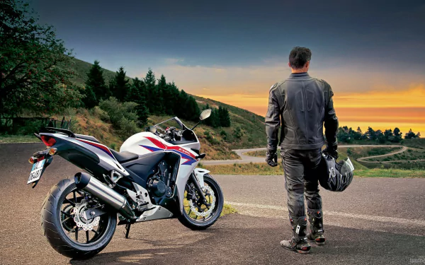 A rider in a black leather suit stands overlooking a winding road at sunset, next to a sleek Honda CBR motorcycle, capturing the spirit of adventure and freedom on two wheels.