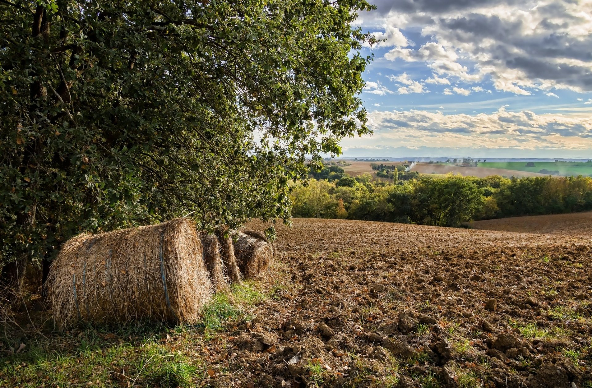 HD PC desktop wallpaper: nature scene with haystack beside a tree, plowed field stretching to green hills under a cloud-dappled blue sky.