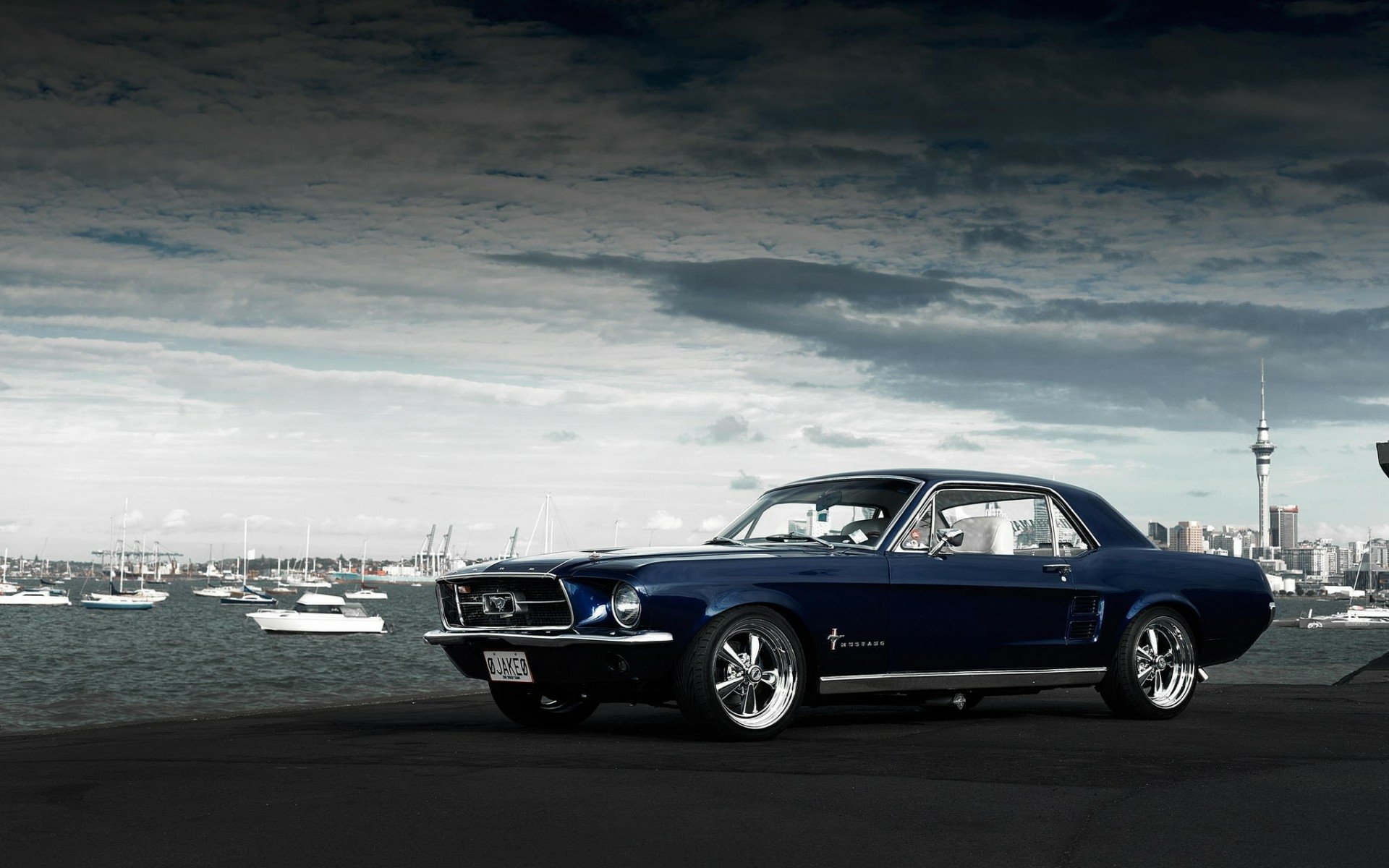 HD desktop wallpaper featuring a classic Ford Mustang parked by a coastal cityscape under a dramatic sky.