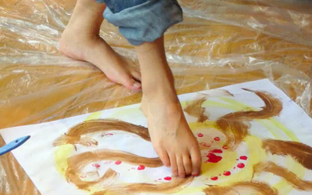 A woman paints with her feet on a large sheet of paper, surrounded by a protective plastic covering, showcasing an engaging and creative foot painting process.