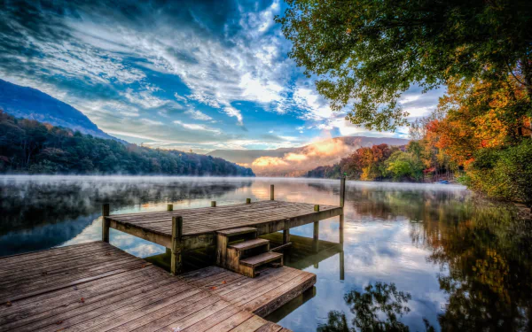 HD PC desktop wallpaper photography: fog over a glassy mountain lake at sunrise, dramatic sky, wooden dock in foreground and colorful autumn shoreline.