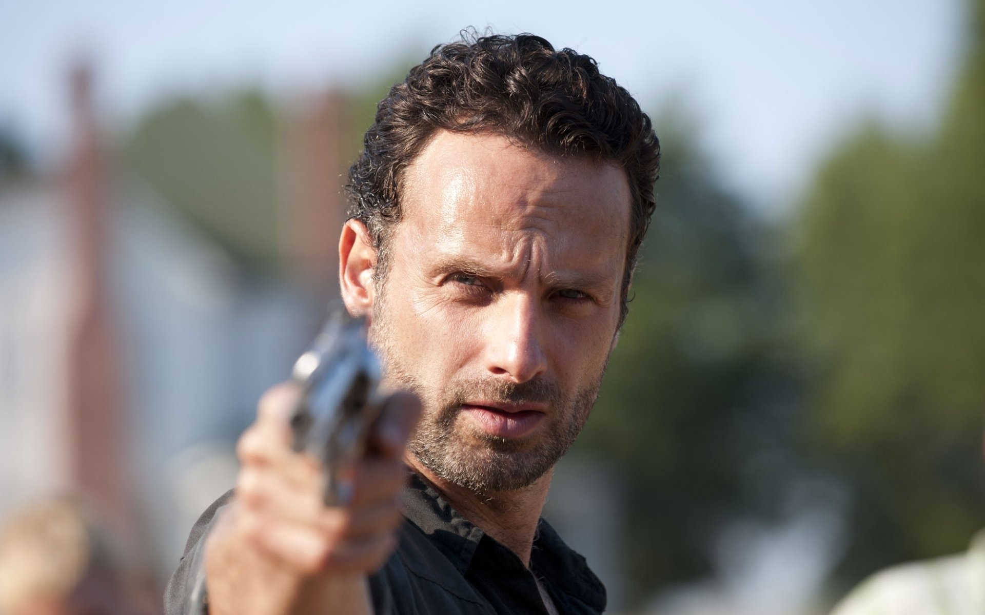 A high-definition wallpaper from the TV show The Walking Dead featuring a character aiming a gun, set against a blurred background of suburban houses.