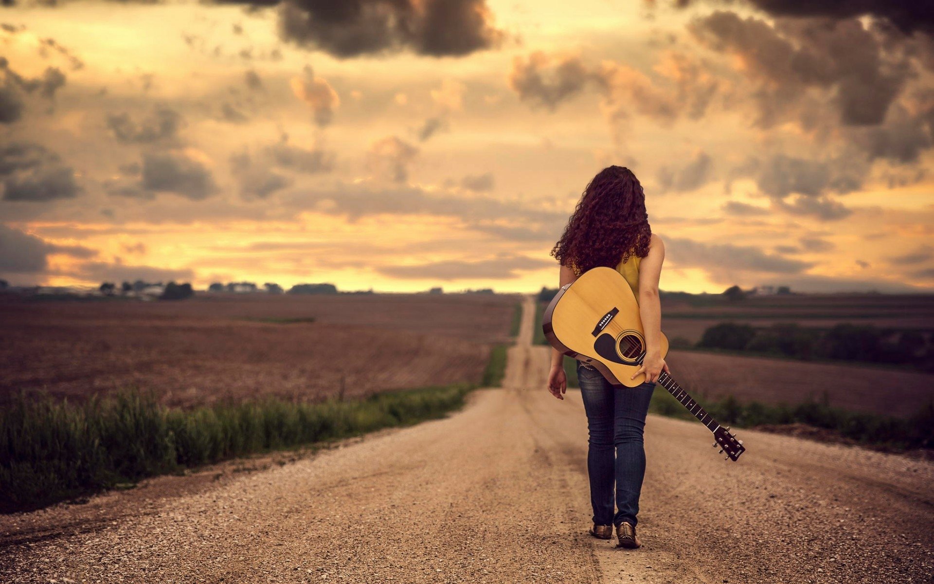 A woman with a guitar walks down a rural road towards a vibrant sunset, creating a serene and inspiring HD desktop wallpaper and background.
