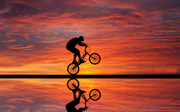 A silhouetted BMX rider performs a trick against a stunning sunset, reflecting on calm waters. Vibrant clouds fill the sky, creating a dynamic sports photography background.