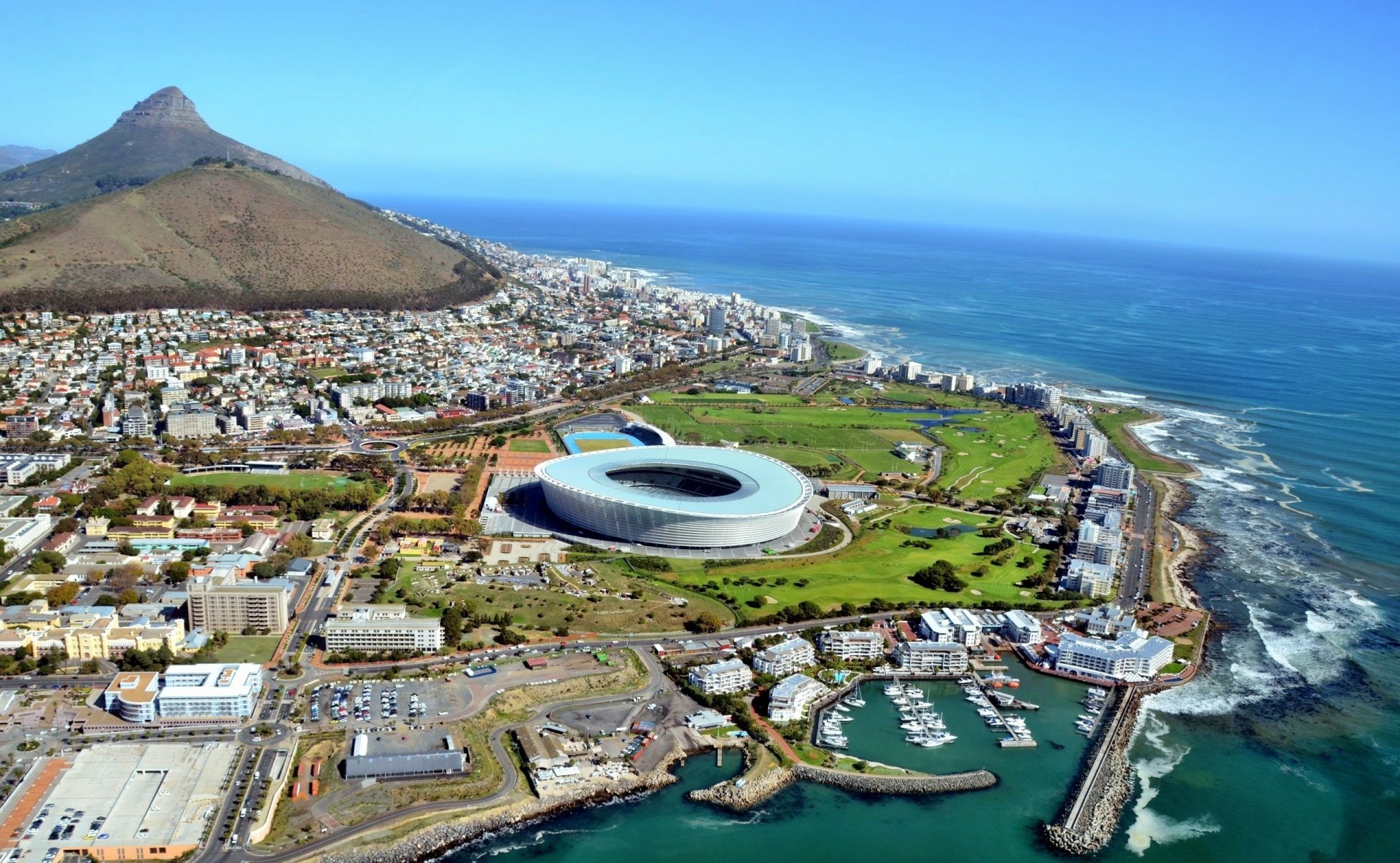 Aerial view of Cape Town, South Africa, showcasing a modern stadium by the seaside, surrounded by lush greenery, mountains, and the ocean's waves, creating a stunning landscape.