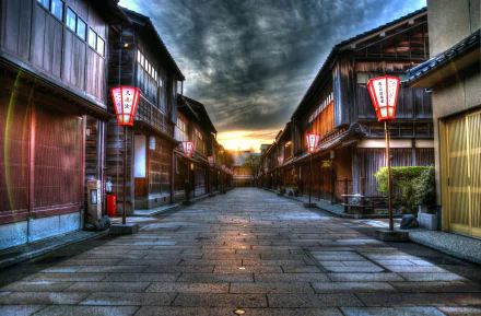 A stunning sunset casts a warm glow over a tranquil street in Kanazawa, Japan, framed by traditional wooden buildings and glowing lanterns, creating a serene man-made landscape.