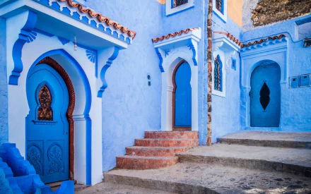 A vibrant HD desktop wallpaper featuring blue-painted houses with arched doorways and steps in the man-made city of Chefchaouen.