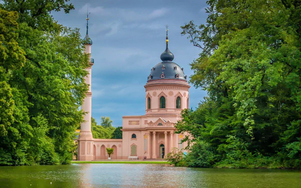 View of the Pink Mosque surrounded by trees and a lake in Schwetzingen, Baden-Württemberg, Germany, showcasing the religious building and its serene landscape.