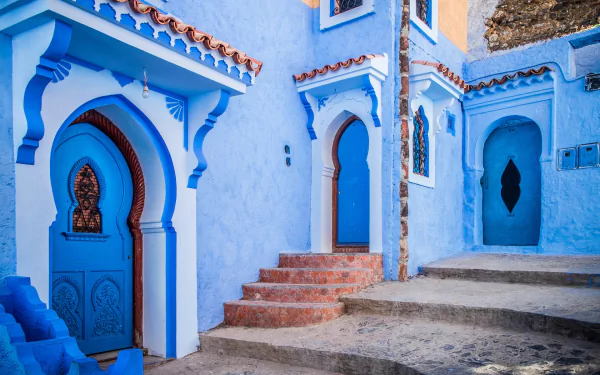 A vibrant HD desktop wallpaper featuring blue-painted houses with arched doorways and steps in the man-made city of Chefchaouen.