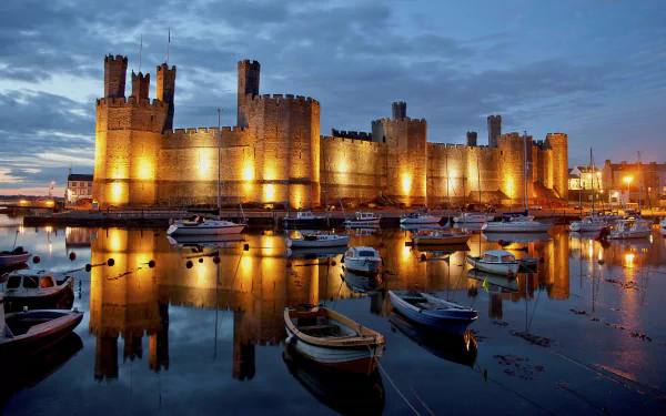 Caernarfon Castle illuminated at night, reflected in calm waters with boats docked nearby, captured in 4K Ultra HD for a striking PC desktop wallpaper.