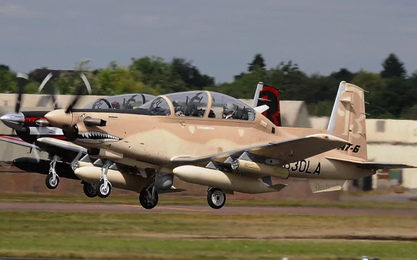 HD PC desktop wallpaper of two military Beechcraft T-6 Texan II trainers in desert camouflage landing on a runway with hangars and trees in the background.