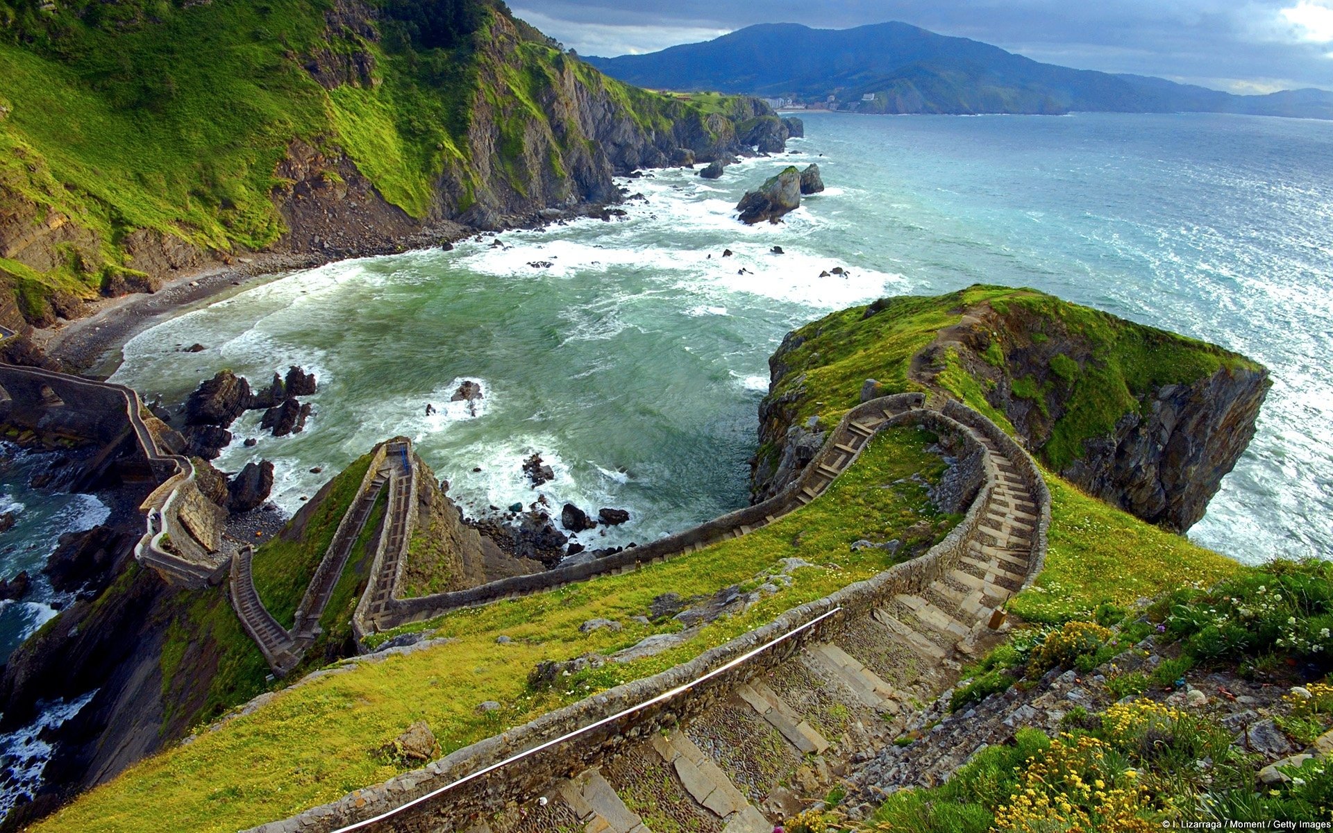 HD wallpaper of a winding man-made staircase along a grassy hill overlooking the sea, with rocky cliffs and mountains in the background.