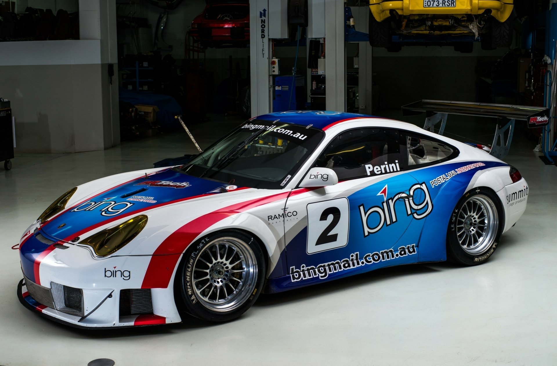 HD desktop wallpaper of a Porsche 911 race car featuring a blue, white, and red livery with sponsor logos, displayed in a garage setting.